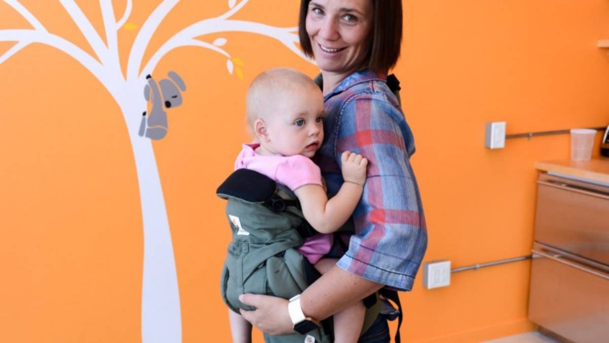 A woman holding a baby in a carrier against an orange wall with a tree mural.
