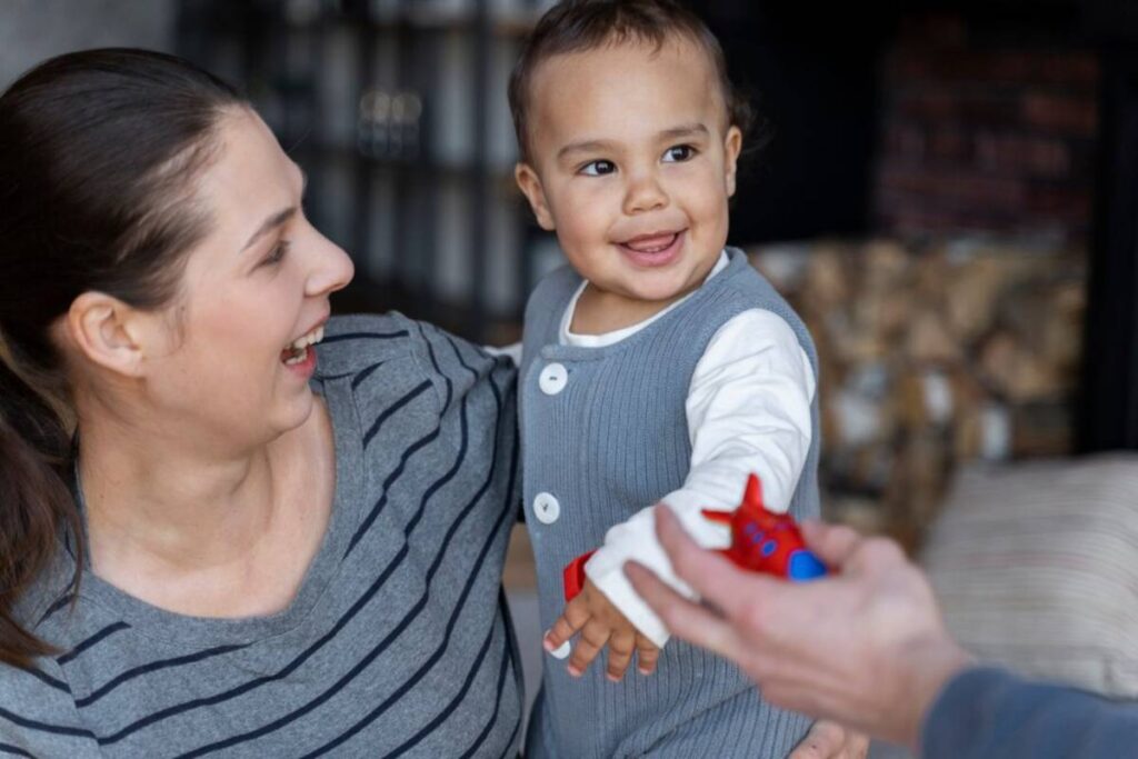 A joyful woman playing with a smiling toddler reaching for a toy airplane.