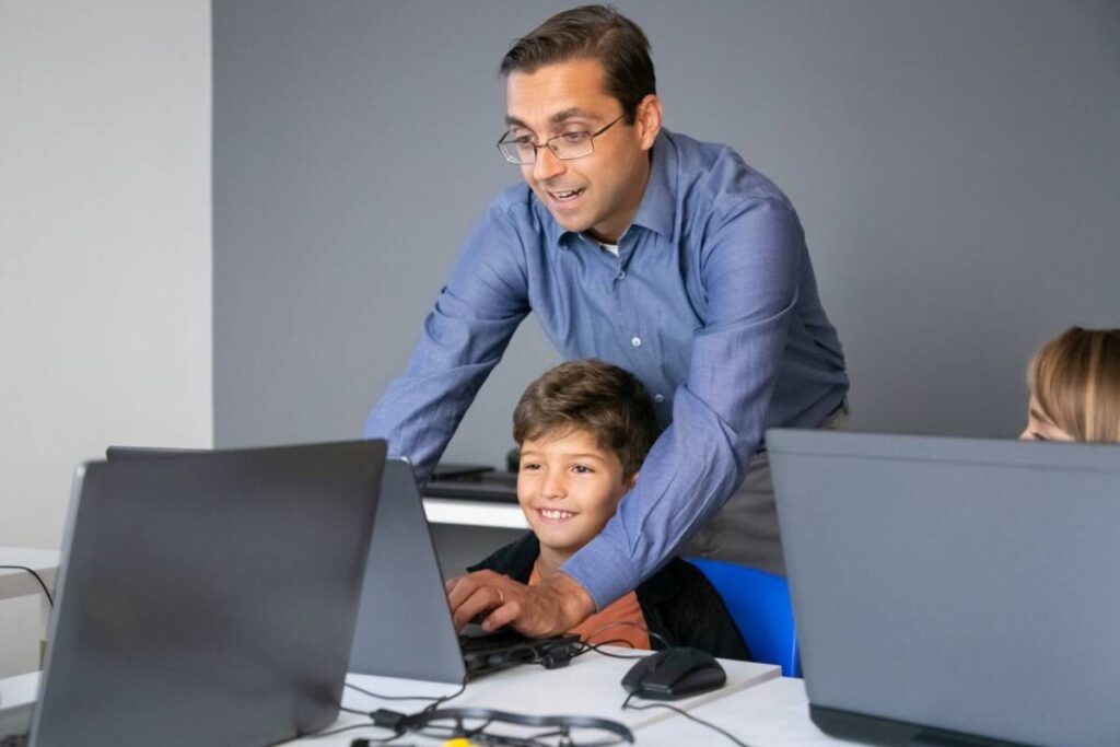 Adult teaching a young boy on a computer in a classroom setting.