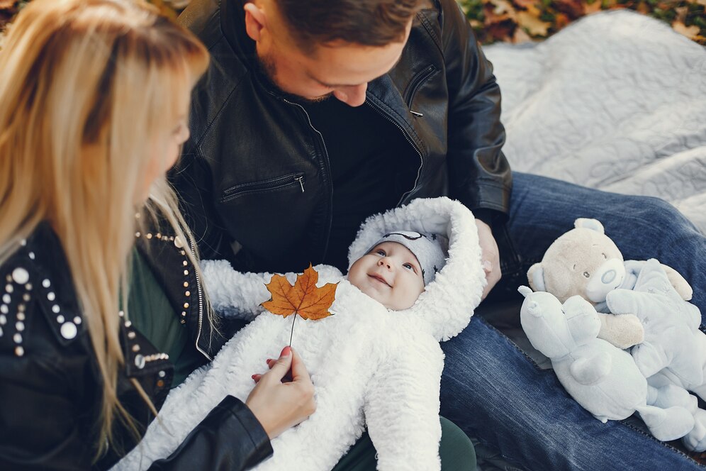A young couple adoring their baby, who is dressed in a cozy white outfit, lying on a blanket outdoors.