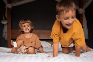 Two young children playing on a bed; one smiling seated child holds a toy, another blurred in motion.