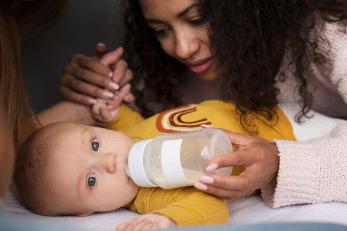 A woman feeding a baby with a bottle while holding the baby's hand.