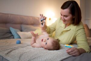 A mother plays with her baby on a blanket, smiling and holding a toy.