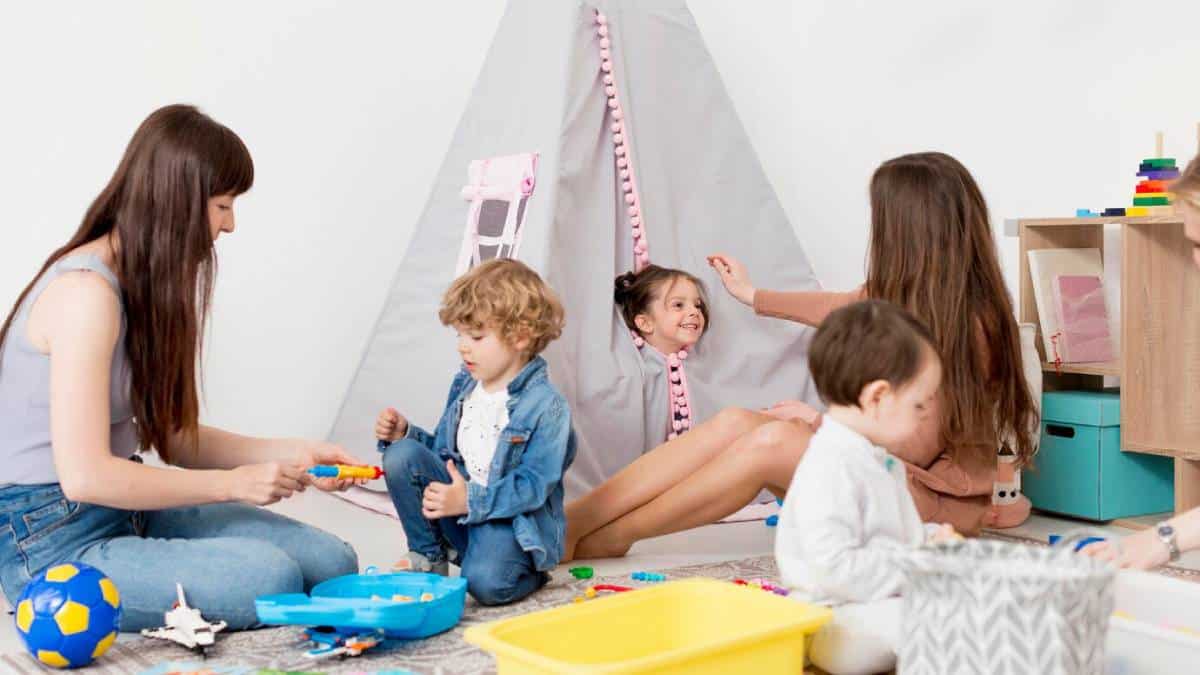 Two women and three young children engaged in playful activities in a bright playroom with a teepee tent.