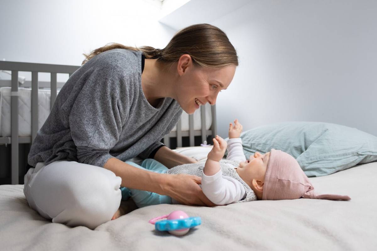 A mother smiling at her baby lying on a bed, with a crib in the background.