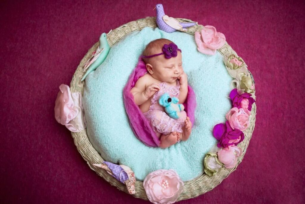 Newborn baby sleeping in a decorated wicker basket with colorful flowers, a soft blue toy, and a purple headband.