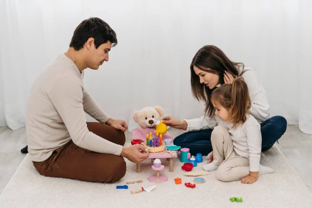 Family playing with toys on the floor, including a young girl, a teddy bear, and colorful blocks.