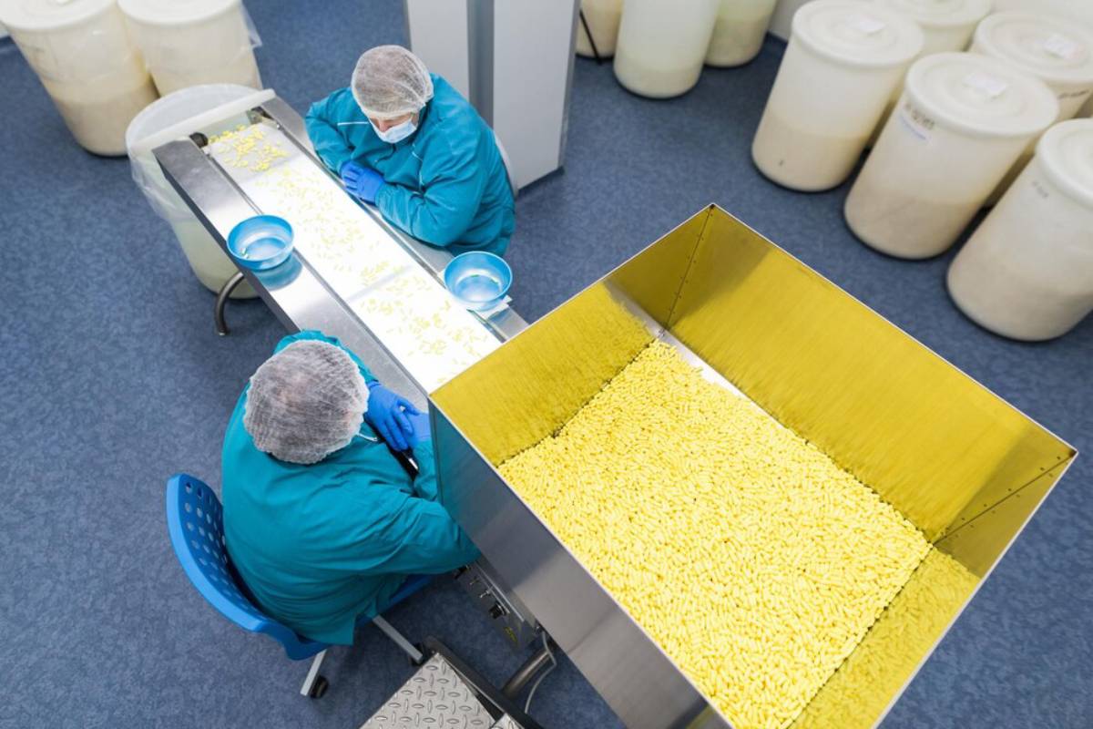 Two workers in protective clothing inspecting pills at a pharmaceutical manufacturing facility.