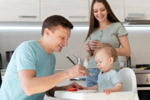 A smiling man feeds a toddler in a high chair while a woman watches, all in a modern kitchen.