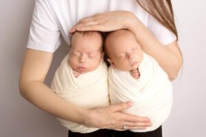 A woman holding twin newborns swaddled in white, sleeping peacefully in her arms.