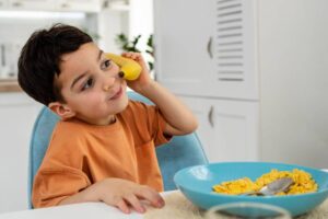 A child sits at a table, holding a banana to their ear, with a blue bowl of cereal in front and a bright kitchen backdrop.