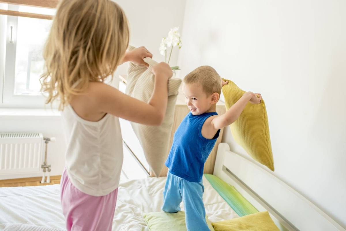 Two young children happily having a pillow fight in a bright bedroom.
