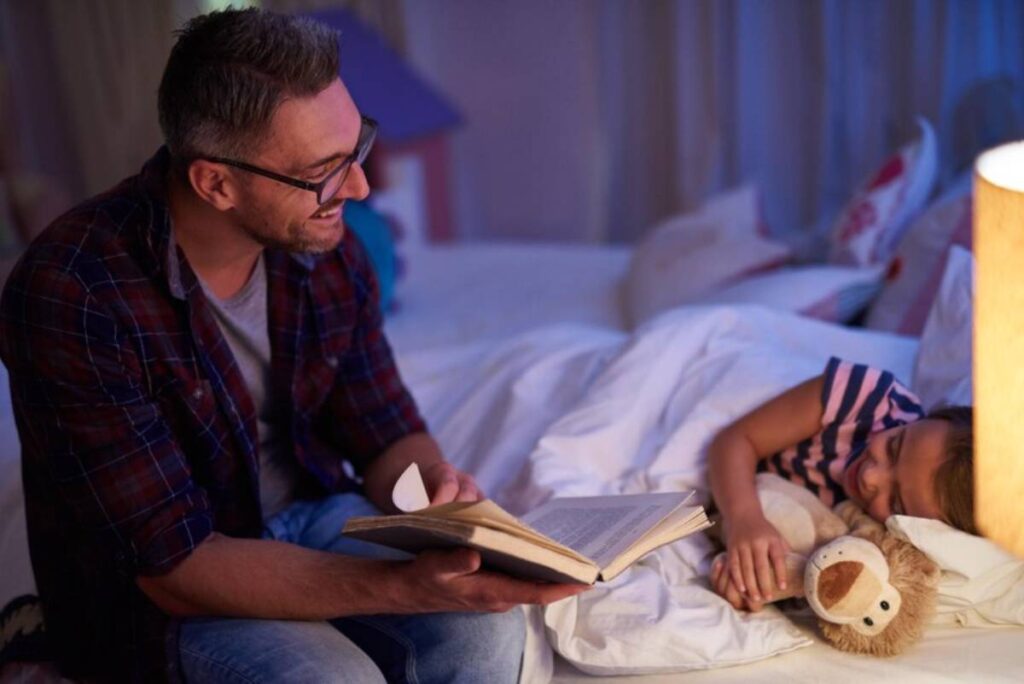 Man in glasses reading a book to a young child in bed beside a teddy bear.