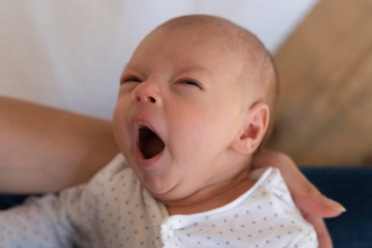 A newborn baby yawning, dressed in a white onesie with tiny stars, held securely in someone's arms.