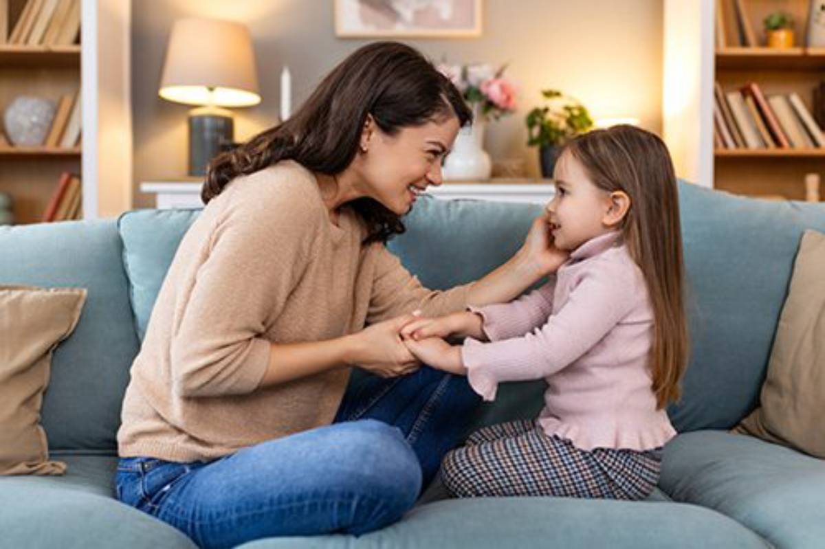 A mother and daughter smiling and holding hands on a couch in a cozy living room.