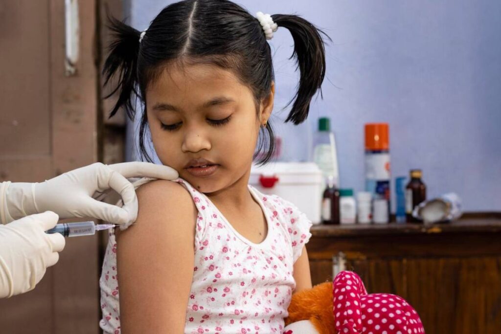 Young girl receiving a vaccination in her arm, holding a stuffed toy, in a medical setting.