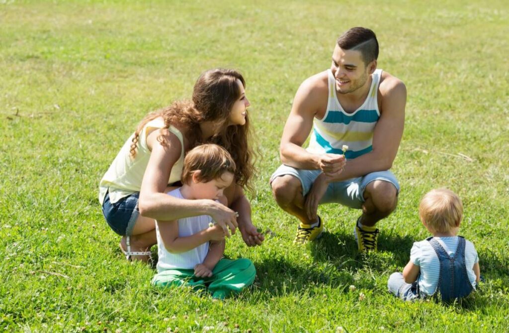 Family of four enjoying a sunny day outdoors in a grassy park, with children playing and parents smiling.
