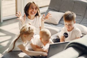 A joyful woman interacting with three young children using a laptop in a sunny living room.