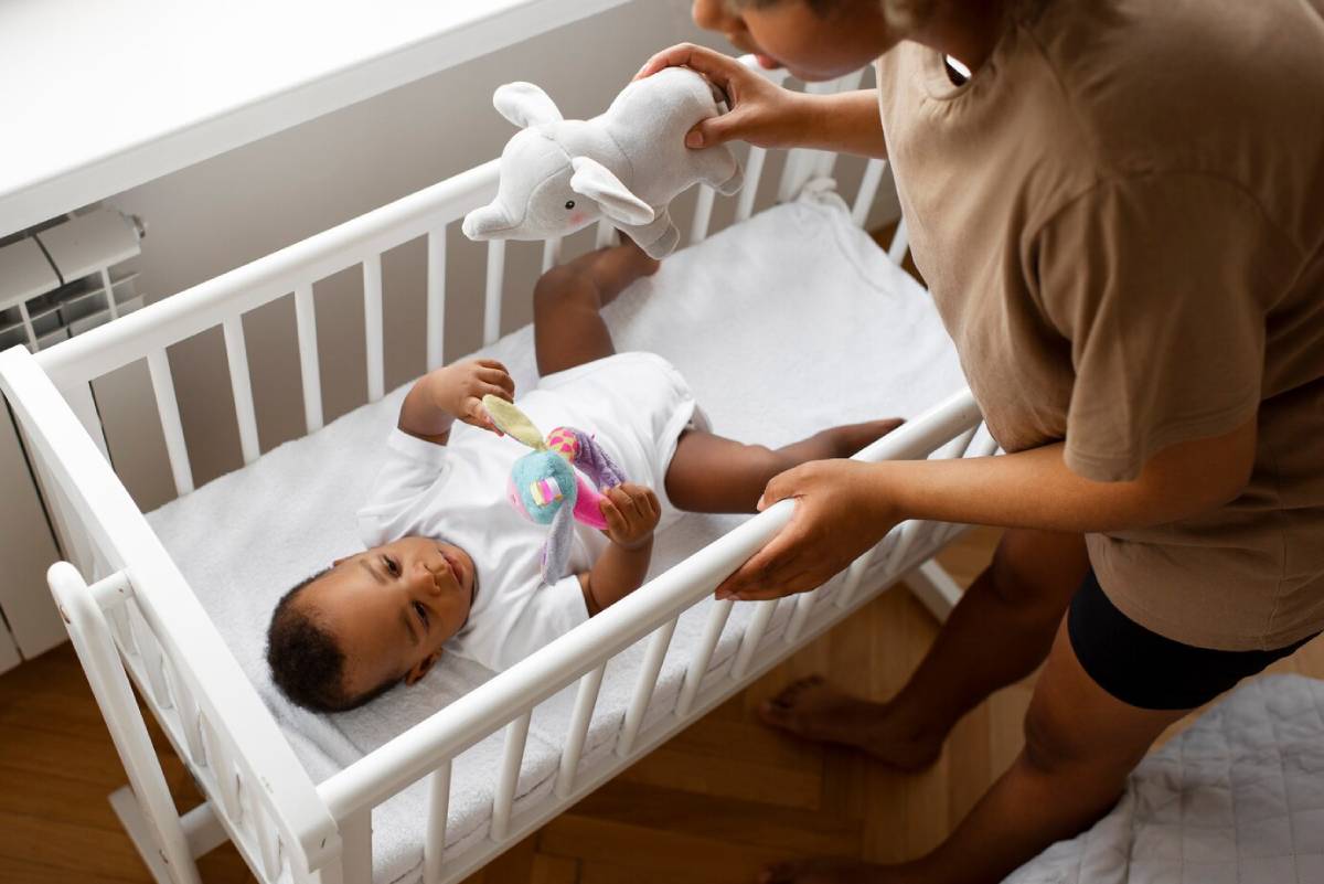 Woman giving a stuffed elephant toy to a smiling baby lying in a white crib.