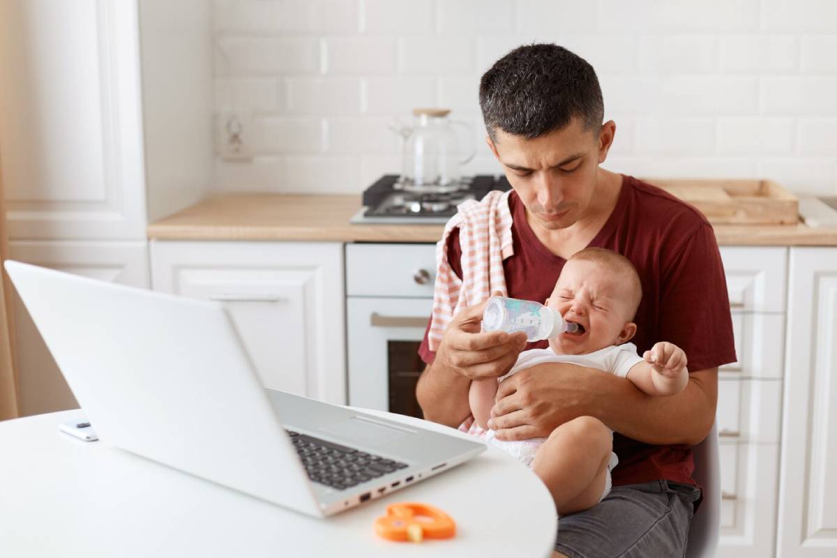 Man feeding crying baby with a bottle while looking at a laptop in a kitchen.