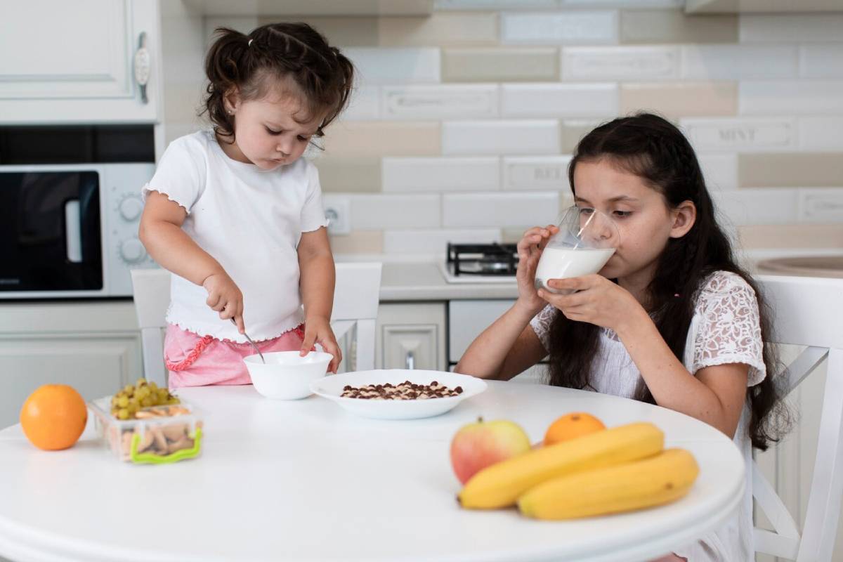 Two young girls eating breakfast in a modern kitchen, with one drinking milk and the other preparing a bowl.