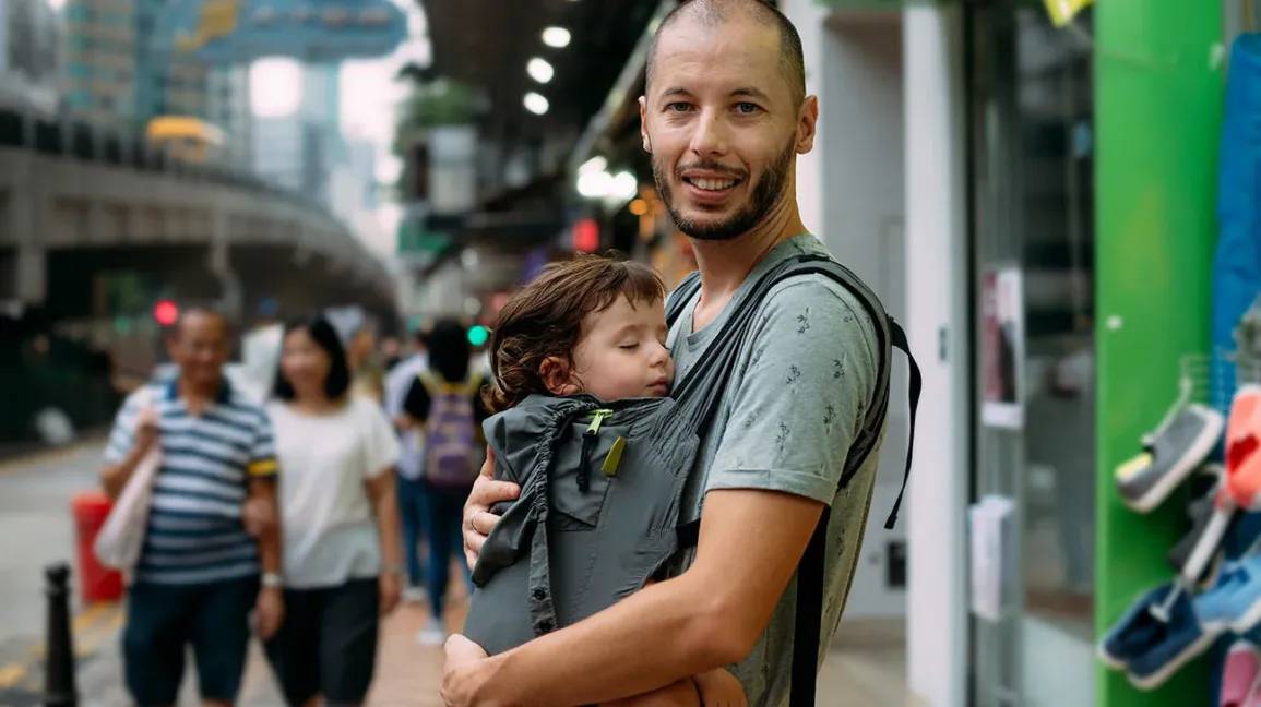 Man with a smiling face carrying a sleeping child in a sling, walking in a busy urban street.