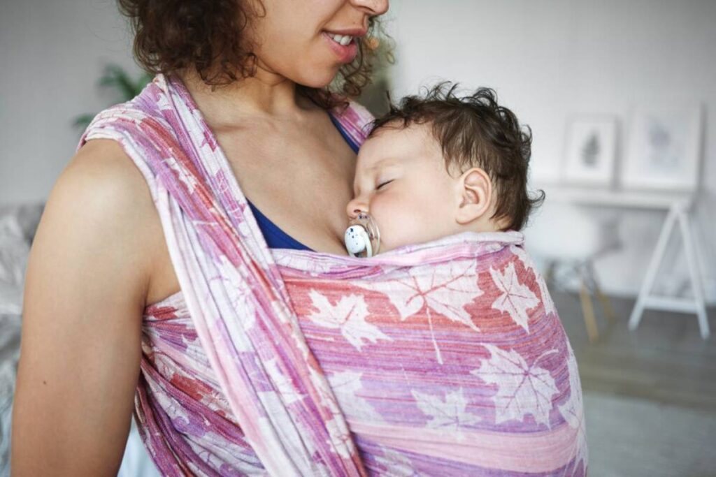 A mother carrying her sleeping baby in a colorful sling, with a pacifier in baby's mouth.