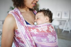 A mother carrying her sleeping baby in a colorful sling, with a pacifier in baby's mouth.