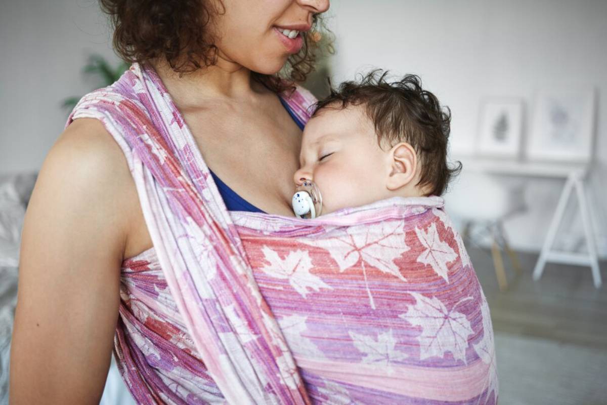 A mother carrying her sleeping baby in a colorful sling, with a pacifier in baby's mouth.