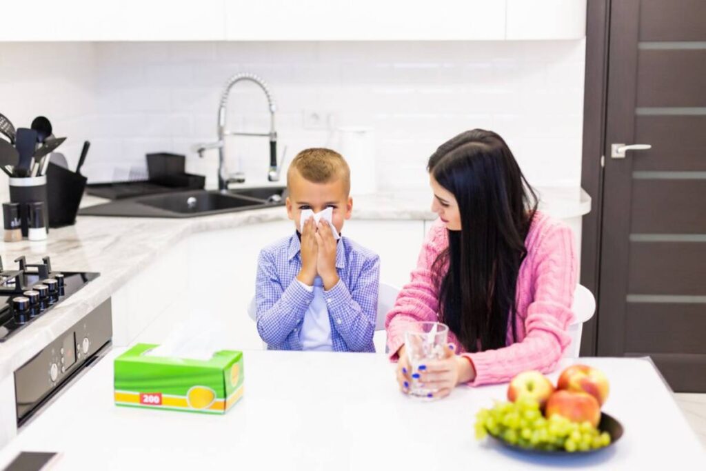 Young boy blowing his nose with tissue while sitting next to a woman in a modern kitchen.
