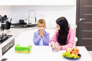 Young boy blowing his nose with tissue while sitting next to a woman in a modern kitchen.