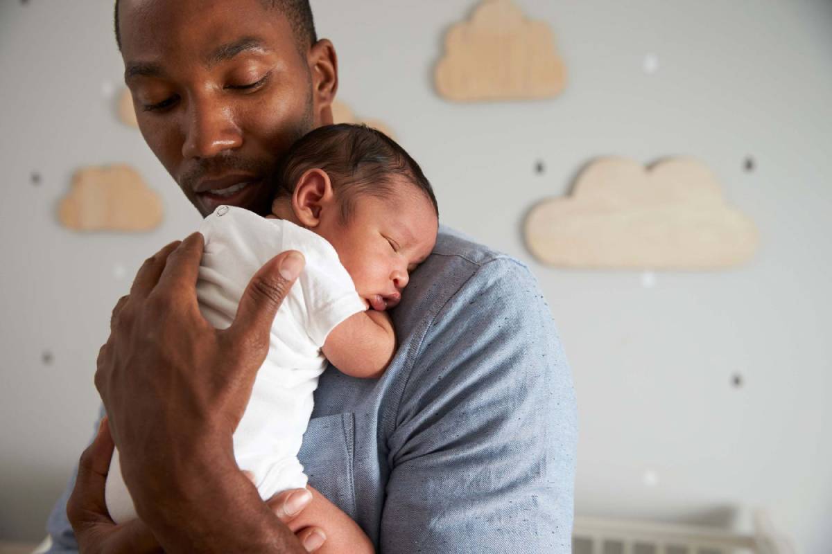 A father gently holds his sleeping newborn in a soothing embrace, against a backdrop of cloud decals on a wall.